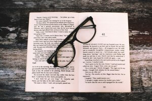 An open book with glasses laying on top, with a wood background behind